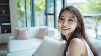 Pretty young smiling Asian woman dressed in white sitting on a couch and looking into camera in modern style living room