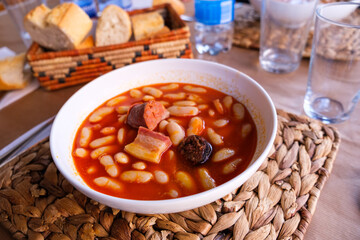 A bowl filled with Fabada asturiana, a traditional Spanish stew made with beans and various meats, sits on a wooden table.