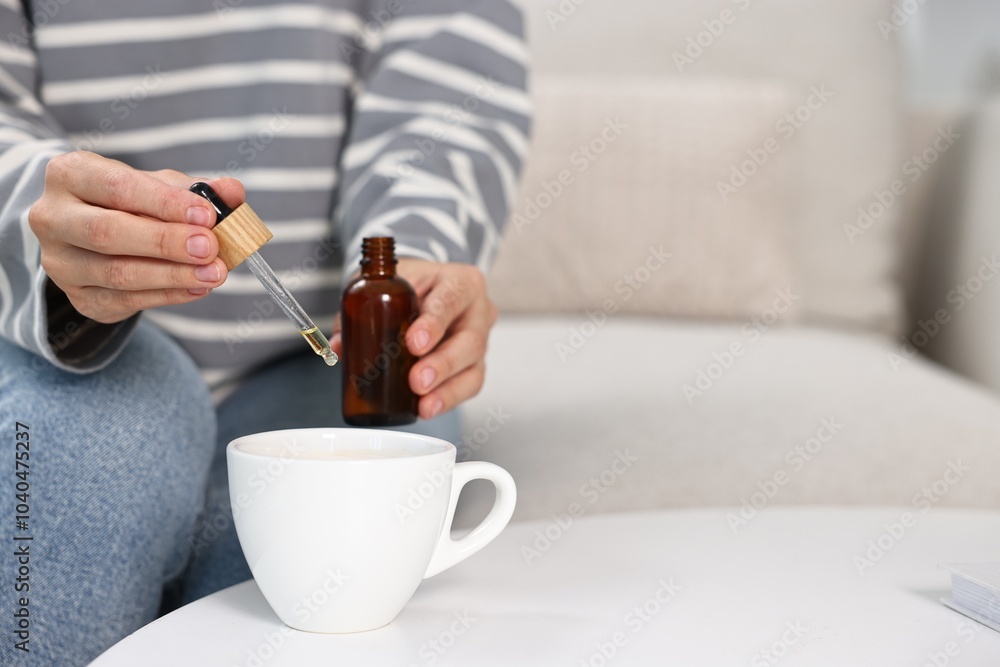 Poster Young woman putting CBD tincture into cup with drink at white table, closeup. Space for text