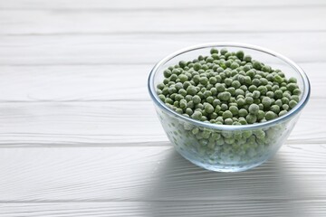 Frozen green peas in glass bowl on white wooden table, space for text