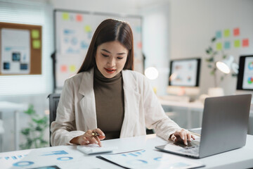 Asian accountant is calculating financial data for a business using a calculator and laptop in her...
