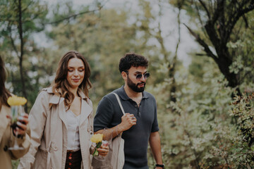 A couple strolls through a scenic forest, holding flowers and wine glasses, enjoying a tranquil autumn day. Their relaxed expressions suggest a moment of connection and nature appreciation.