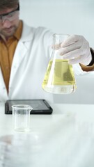 Man scientist wearing white protective gloves, and glasses is holding a yellow chemical solution inside an Erlenmeyer flask in a laboratory, vertical portrait