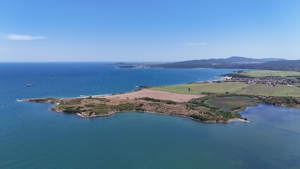 A stunning aerial view of a coastal landscape featuring a serene bay, vast green fields, and distant mountains under a clear blue sky. The image captures a peaceful seaside area with a few ships