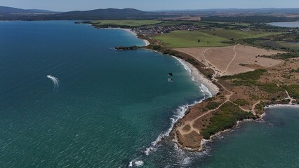 A stunning aerial view of a coastal landscape featuring a serene bay, vast green fields, and distant mountains under a clear blue sky. The image captures a peaceful seaside area with a few ships
