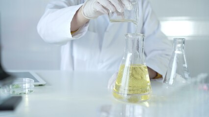 A scientist, wearing a lab coat and white protective gloves, is pouring a yellow oily liquid from one beaker to another in laboratory, close up. Medicine and science concept