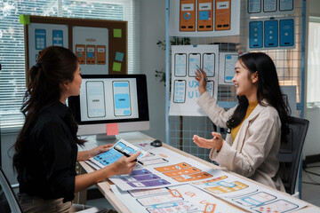 Two asian female uiux designers are discussing over mobile app interface wireframe design at their office desk