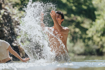 Fototapeta premium A young man energetically splashes water in a natural outdoor environment, expressing joy and freedom in the sunshine. The playful scene captures the essence of summer fun and relaxation.