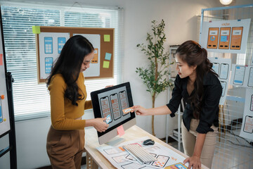 Two asian female uiux designers are discussing over mobile app interface wireframe design on desk...