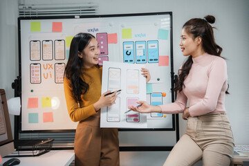 Two happy asian female uiux designers discussing over mobile application interface wireframe design...