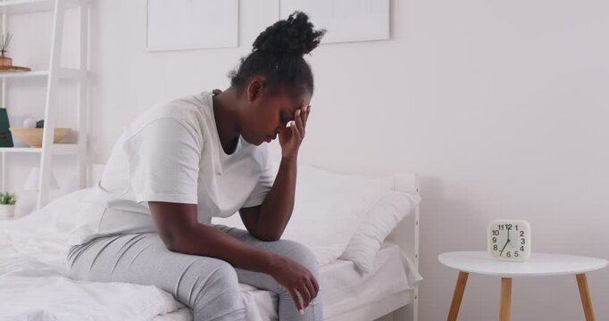 Exhausted young African American woman sitting on bed early in morning, holding head with her hand. Tired black girl feeling weary and unrefreshed after waking up at 7 AM and poor night sleep at home.