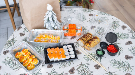 Delivery of sushi and rolls in food containers on the table in the festive interior of the kitchen of the house with a white Christmas tree for Christmas and New year. Food delivery for the holidays