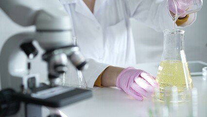 Fototapeta premium A scientist, wearing a lab coat and pink protective gloves, is pouring a yellow oily liquid from one beaker to another near microscope in laboratory, close up. Medicine and science