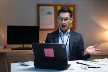 Shocked asian businessman is looking at his laptop with a confused expression, raising his hands in frustration while working late at night in the office