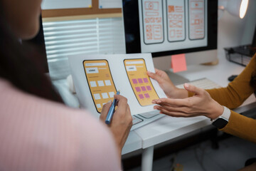 Two ui designers are discussing the wireframe design of a mobile application, holding paper...