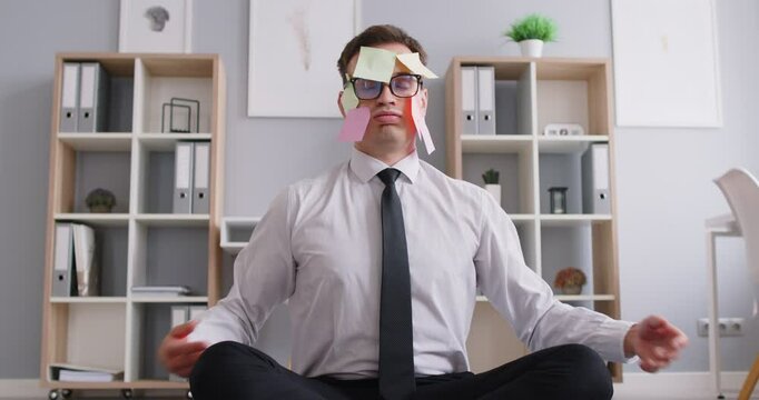 Calming down in stressful situation, office man with sticky notes stuck on face sitting cross-legged in easy yoga namaste pose at workplace, overworked, meditating on break time to relax, breathing 