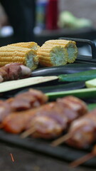 Close-up of marinated meat skewers grilling on a campfire with corn. The scene focuses on the texture and details of the food, evoking a sense of outdoor culinary experience