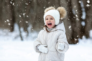 Happy laughing little girl in warm white jacket and white knitted hat has fun and playing with a snow in winter park or forest. Children outdoor activities concept. Happy family time