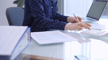 A female accountant with blue dotted blousy is using a laptop computer and calculator to calculate...
