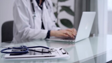 Tablet computer and blue stethoscope are on the glass table while doctor woman is using laptop computer on the glass table. Medicine concept