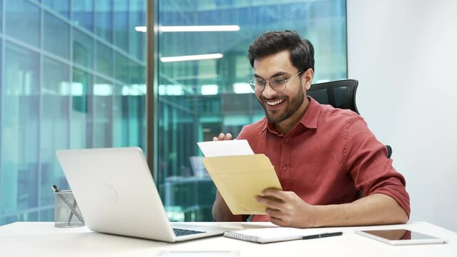 Joyful businessman reading letter with great news sitting at workplace in business office. Excited bearded worker is celebrating success, happy to receive good notification after opening the envelope
