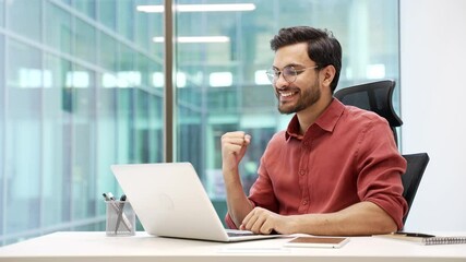 Satisfied happy handsome businessman received great news on laptop while sitting at workplace in business office. Smiling glad bearded entrepreneur reads a positive good message on the computer
