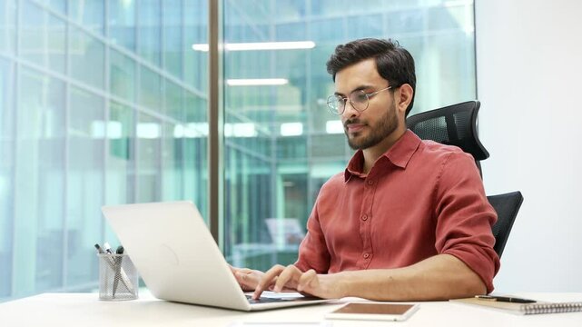 Focused male professional at workplace typing on laptop in modern office desk. Businessman wearing glasses engaged in work, focusing on business tasks. Handsome bearded male worker works on a computer