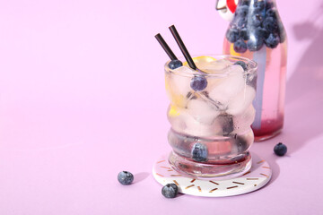 Glass and bottle of fresh blueberry lemonade on lilac background
