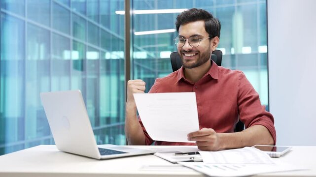 Happy financier satisfied with financial results sitting at workplace in business office. Smiling handsome bearded businessman looks at documents and computer and celebrates success looking at camera