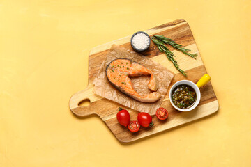 Wooden board of tasty salmon steak with tomatoes and sauce on yellow background