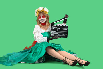 Young woman with painted skull on her face and clapperboard sitting against green background. Halloween celebration