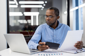 Mature african american businessman reviewing tax forms in office, holding calculator. Uses smartphone for calculations alongside laptop. Scene conveys focus, analysis, financial management.