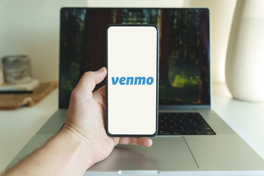 Venmo app logo on cellphone screen. Person holding Venmo application on smartphone in laptop background