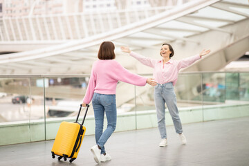 Two friends embrace joyfully at a contemporary airport terminal. One friend arrives with a bright yellow suitcase, excited to reconnect after time apart.