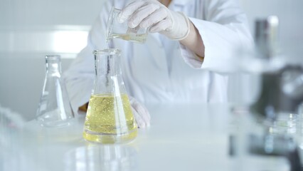 A scientist, wearing a lab coat and white protective gloves, is pouring a yellow oily liquid from one beaker to another in laboratory, close up. Medicine and science concept