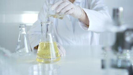 A scientist, wearing a lab coat and white protective gloves, is pouring a yellow oily liquid from one beaker to another in laboratory, close up. Medicine and science concept