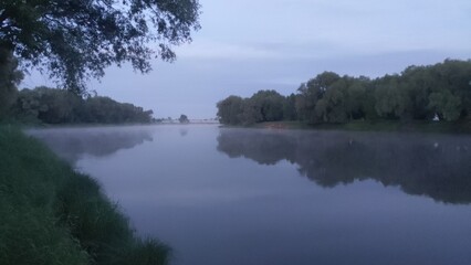 On a summer morning, the sun had not yet risen in a cloudy sky. Over the water is a fog. On the calm water reflections. The beauty of nature. Shrubs and trees grow on the river banks. Twilight