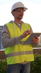 Obraz premium Male engineer with white helmet and safety vest is taking notes on a digital tablet while inspecting a construction site at sunrise in early morning, vertical front view. Architecture and engineering