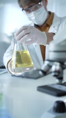 Man scientist researcher, wearing white protective gloves, mask and glasses, is holding a yellow chemical solution inside Erlenmeyer flask in a laboratory, vertical portrait view. Science and medicine
