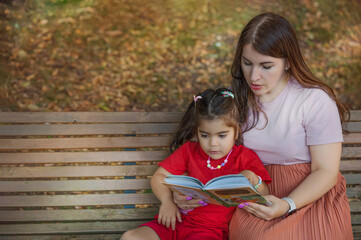 A young mother and daughter are reading a book on a bench in the park.