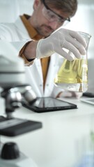 Man scientist, wearing white gloves and protective glasses, is holding a yellow chemical solution...