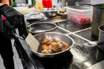 A close-up of a gloved hand cooking meat in a stainless steel pan on a stove. The blurred kitchen background features various kitchenware and ingredients, creating a professional culinary atmosphere.