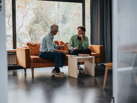 An older and younger man sitting on a sofa having an engaging conversation in a modern interior setting. The scene conveys a sense of mentorship and sharing ideas.