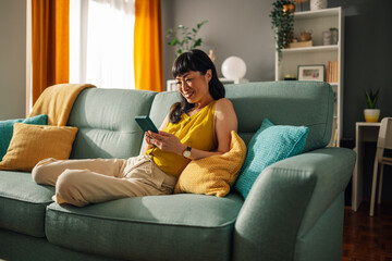 Asian woman in yellow top relaxes on teal sofa, using smartphone, cozy room.