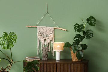 Chest of drawers with glowing lamp, Monstera houseplant, human figure and books near green wall, closeup