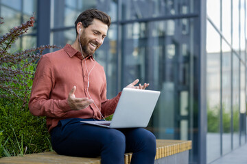 Businessman using laptop for digital meeting outdoors. Smiling in casual attire with earphones,...