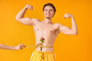Fototapeta premium Female hand pulling tape measure around young man's waist with salad on yellow background. Weight loss concept