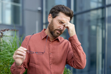 Man experiencing fatigue holds glasses, potentially suffering headache or eye strain outdoors....