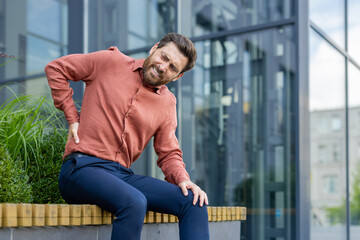 Person in orange shirt sitting outdoors with visible back pain expression. Urban setting with...
