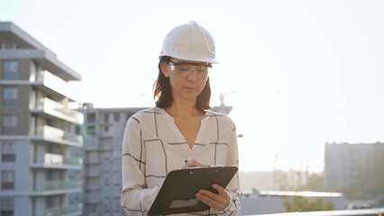 Woman construction engineer wearing white checked blouse and hard hat is making notes on a clipboard while inspecting a building site at sunset, front view. Architecture and engineering concepts